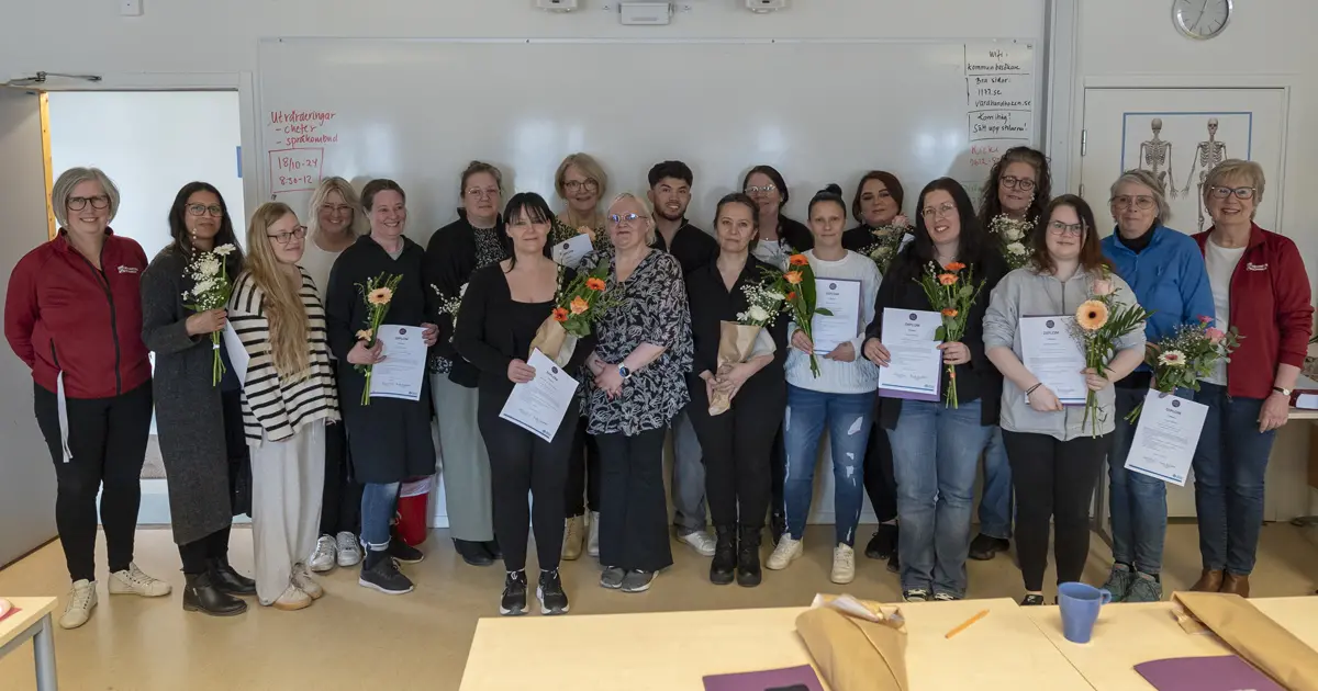 Flera språkombud och deras lärare står i ett klassrum med diplom och blommor i sina händer.
