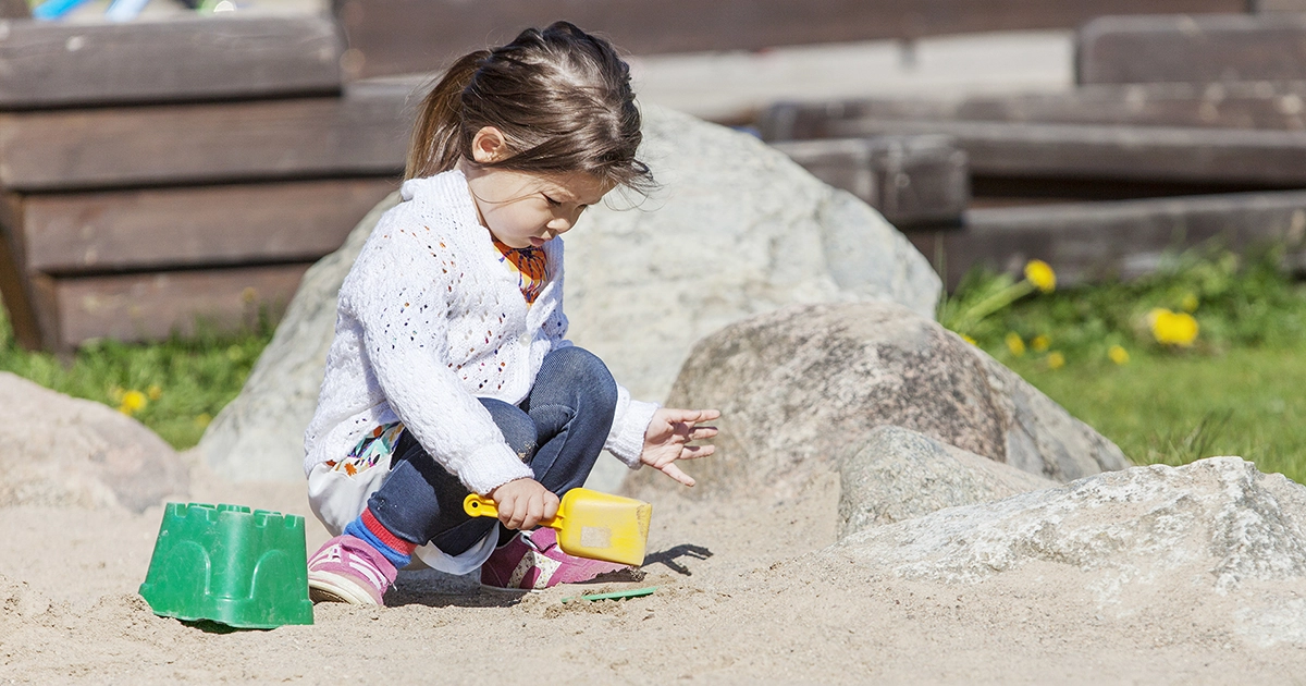 En liten flicka klädd i jeans och kofta sitter i en sandlåda. Hon har en gul spade i handen och ska gräva.