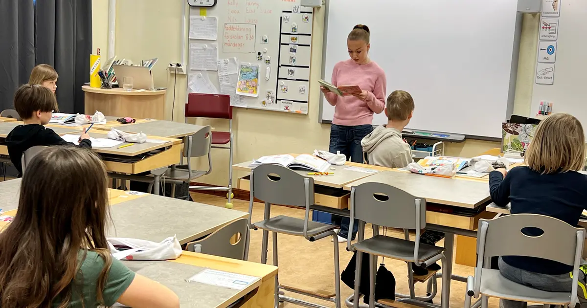 Matilda har en rosa tröja och blå jeans. Hon står längst fram i klassrummet och läser ur en bok. I bänkarna sitter elever och lyssnar. 