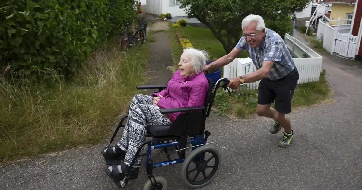 En man skjuter en rullstol uppför en backe. I rullstolen sitter en äldre kvinna.