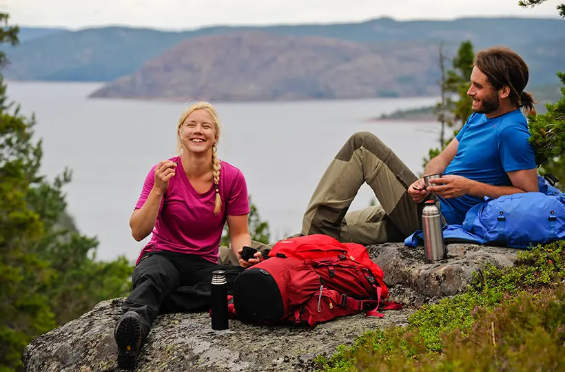 En ung man och en ung kvinna sitter på en klippa i Höga Kusten. De har en ryggsäck och en termos bredvid sig.