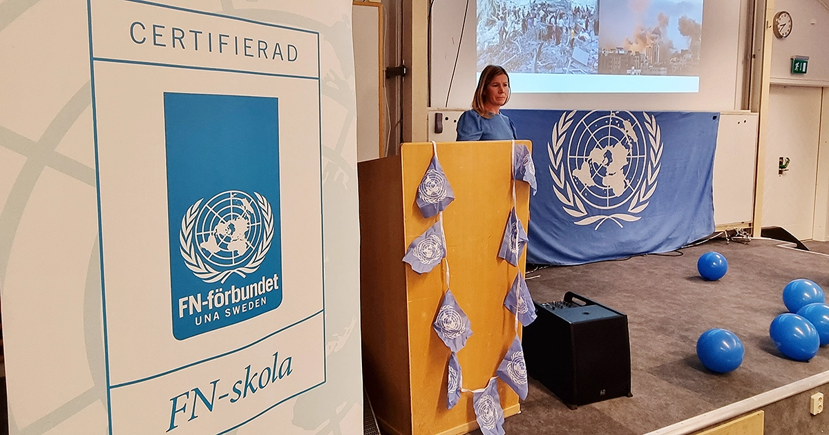 En rollup med budskapet att Räddningsgymnasiet är FN-skola och läraren Elin Gradin som inviger dagen. Talarstolen är smyckad med blå ballonger.