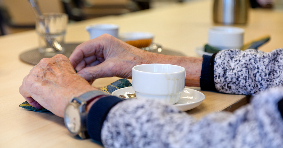 En äldre persons armar och händer vilar på ett bord. Framför finns en kaffekopp.