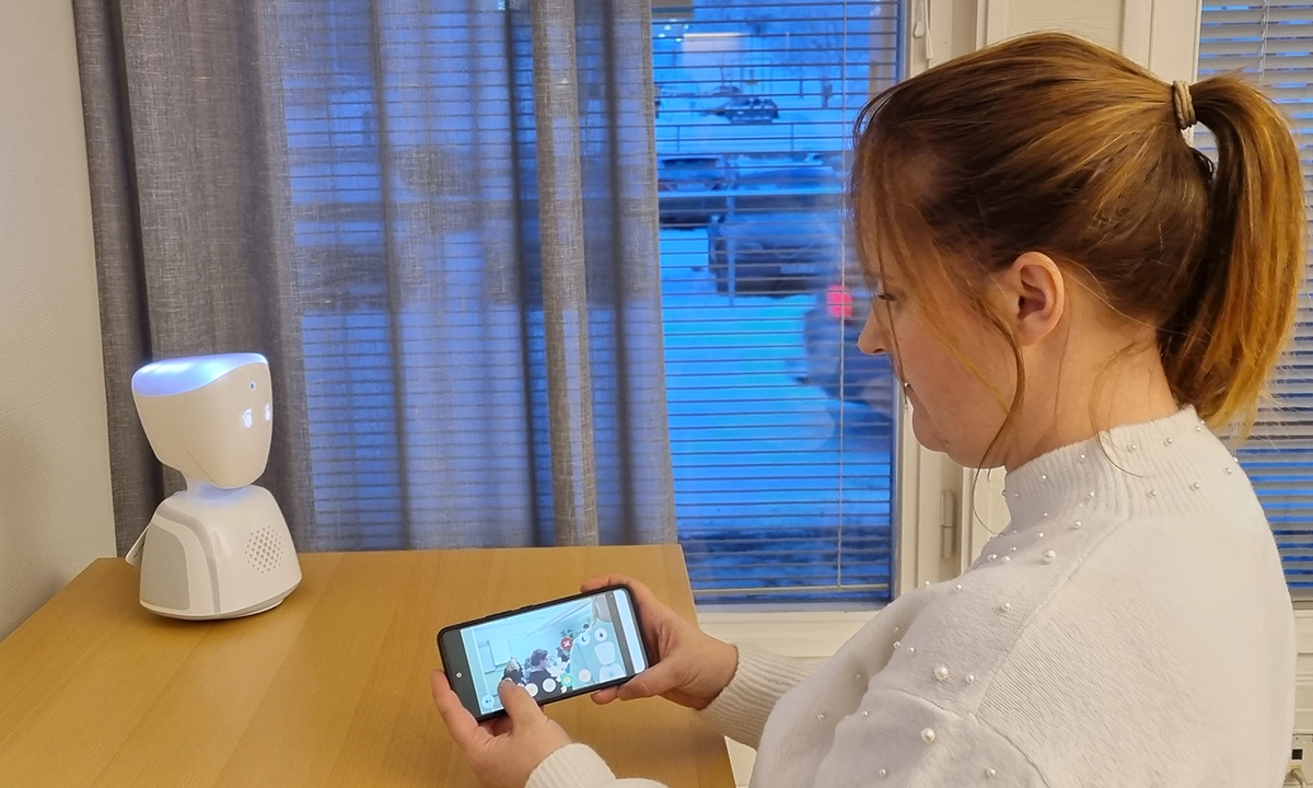 LIsa Karlsson har vit tröja och håret uppsatt i hästsvans. Roboten står på ett bord och Lisa testkör den med sin mobil.