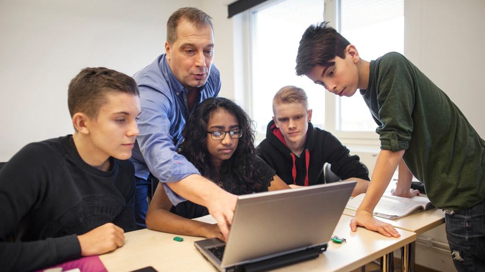 Ett antal elever sitter vid ett bord och tittar i en dator. En manlig lärare och med. Han pekar på något i datorn.